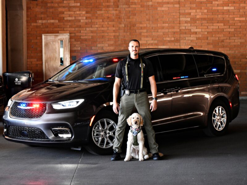 A police van is in the background with an officer standing in a navy shirt and green pants with his partner, a golden doodle named Finley sitting at the center. Both have police patches on their chest.