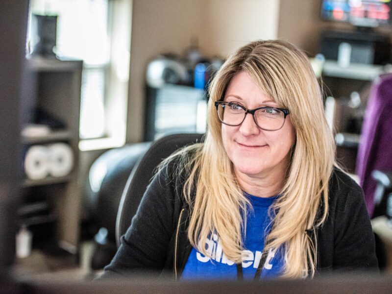 A female dispatcher with glasses smiles at her computer screen with office equipment in the background.